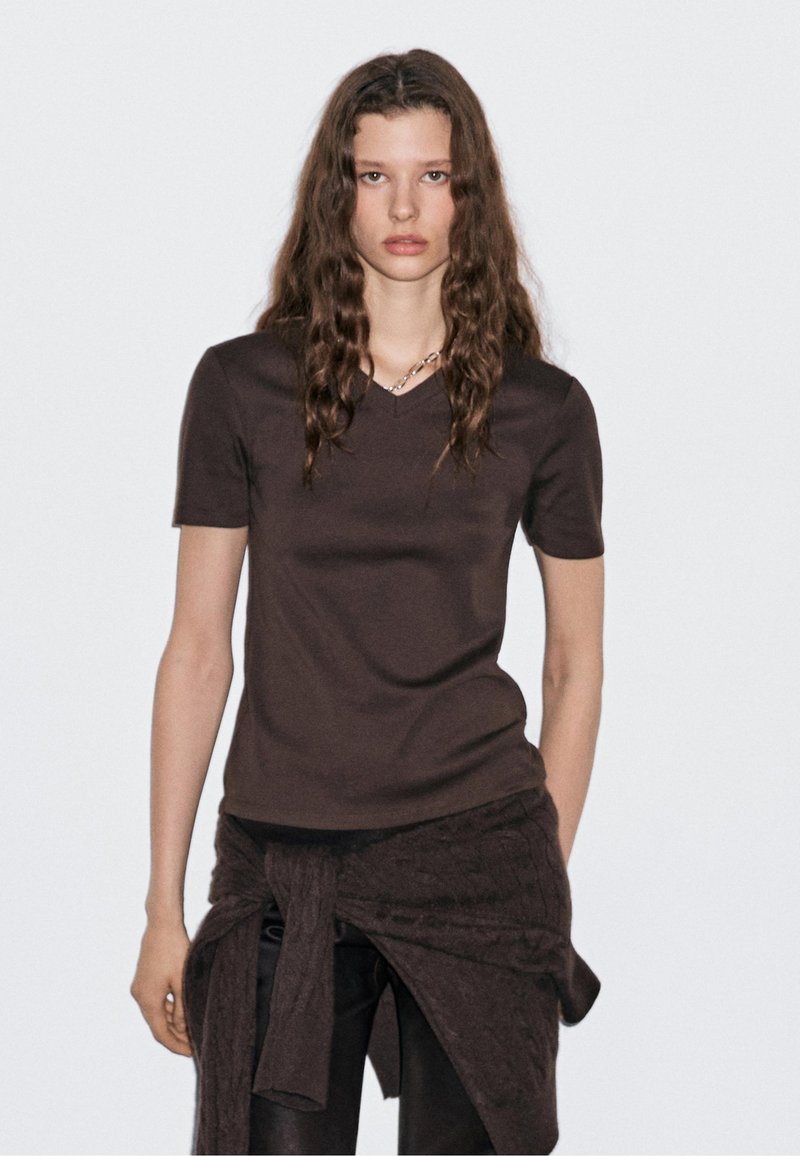 T-shirt marrone a V con maniche corte, realizzato in tessuto liscio, indossato con un avvolgente scuro e texture. Design semplice con una forma aderente.