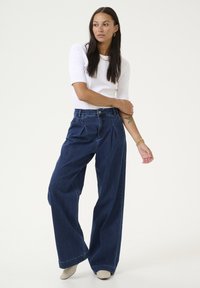 Leveled denim-housut tummansinisenä, joissa on korkea vyötärö, poimutettu etuosa ja ommeltu yksityiskohtia. Yhdistetty tiukkaan valkoiseen puseroon.