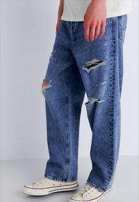 Jeans en denim bleu avec des détails usés et de grandes déchirures, affichant une coupe ample et un design à jambe droite, associés à des baskets blanches.
