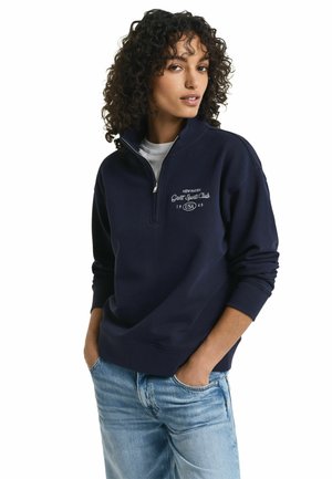 Sweat-shirt zippé navy avec col haut, présentant une broderie sur le devant et des poignets côtelés. Porté avec un jean bleu clair, affichant une coupe décontractée.