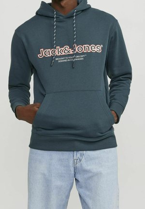 Osoba nosząca ciemnozieloną bluzę z kapturem Jack & Jones z przednimi kieszeniami i jasnoniebieskie dżinsy, ręce częściowo w kieszeniach na jednolitym tle.