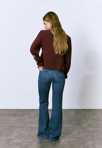 Blusa burdeos texturizada con mangas acampanadas, combinada con jeans acampanados azules que tienen bolsillos traseros. El cabello largo y lacio cae sobre el hombro.