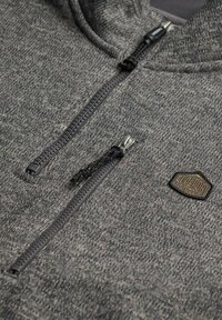 Close-up van een grijs текстuur zip-up kledingstuk met twee zwarte ritsen en een klein hexagonaal logo patch.