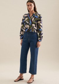 Blusa a maniche lunghe con un motivo astratto colorato, caratterizzata da verde, blu e rosso su uno sfondo crema; abbinata a jeans blu a vita alta.