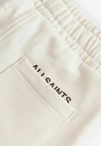 Pantaloni corti jogger color crema con vita elasticizzata e tasca cucita. Presenta il logo 'ALL SAINTS' in stampa scura sulla tasca posteriore.