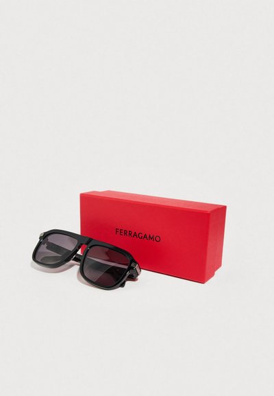 Μαύρα ορθογώνια γυαλιά ηλίου με σκουρόχρωμους φακούς, που διαθέτουν ασημένιες λεπτομέρειες στις μεντεσέδες, δίπλα σε ένα κόκκινο ορθογώνιο κουτί με την ετικέτα "FERRAGAMO".