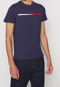 Námořnické bavlněné tričko s kulatým výstřihem a krátkými rukávy; obsahuje horizontální pruhovaný design s nápisem "TOMMY JEANS" v červené, bílé a černé barvě.