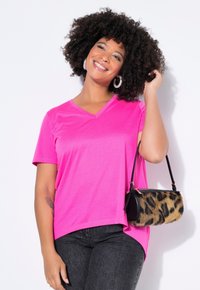 Helles pinkes V-Ausschnitt-T-Shirt mit kurzen Ärmeln, kombiniert mit schwarzen Jeans. Hält eine Handtasche mit Leopardenmuster und schwarzem Pelz. Lockiges Haar ist gestylt.