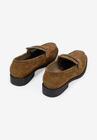 Mocasines de ante marrón con punta redonda, suela plana de goma negra y detalles cosidos. Presentan una solapa plegable en la parte superior.