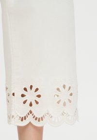 Λευκή φούστα με κυματιστό τελείωμα και κεντήματα με λουλουδάτο μοτίβο, που αποκαλύπτουν μέρος ενός ποδιού πάνω σε απλό φόντο.