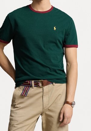 Homme portant un t-shirt vert foncé avec des garnitures rouges, un pantalon beige, une ceinture rayée, et une montre en cuir marron au poignet gauche.