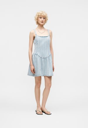 Młoda kobieta z kręconymi blond włosami, ubrana w niebiesko-białą, paskowaną sukienkę bez rękawów i beżowe sandały, stojąca na jednolitym białym tle.