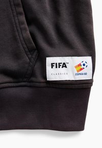 Μαύρο ύφασμα με ραμμένη λευκή ετικέτα που δείχνει «FIFA Classics» και ένα λογότυπο με μια ποδοσφαιρική μπάλα και τα χρώματα της ισπανικής σημαίας με κείμενο «España 82».
