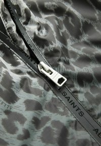 Grått tyg med ett subtilt leopardsmönster, med en silverfärgad dragkedja och varumärke längs dragkedjans kant. Slät textur, lättviktsdesign.