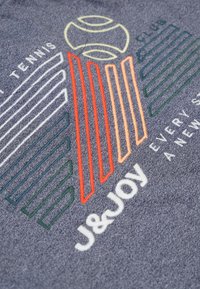 Donkergrijze stof met kleurrijke borduursels in groen, rood en oranje, en witte tekst met de woorden "J&JOY" en "Tennis Club."