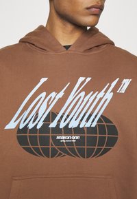 Hnedá mikina s kapucňou, ktorá má veľký grafický motív s nápisom "Lost Youth" v modrej farbe a čiernym globe designom. Obsahuje predné vrecko a kapucňu so šnúrkou.
