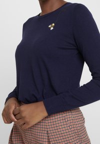 Navyblå stickad tröja med rundad halsringning, prydd med en broderad bi-detalj på vänster axel. Bärs med en rutig kjol.