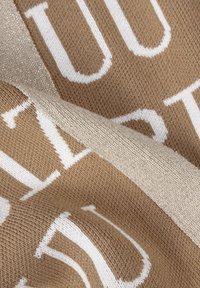 Gebreide stof in warm beige met witte tekst, met een subtiele glans in de textuur. Het ontwerp bevat vetgedrukte, contrasterende letters.