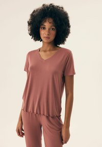 Koszulka z dekoltem w serek i krótkim rękawem, wykonana z jasnoróżowego materiału, o luźnym kroju i falbaniastym dole. Gładka tekstura, odpowiednia do noszenia w domu lub na co dzień.