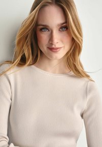 Jasnobeżowy, prążkowany top z długim rękawem i okrągłym dekoltem. Miękka tekstura, dopasowany fason i minimalistyczne detale, w połączeniu z naturalnym makijażem.