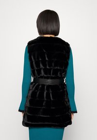 Svart fuskpälsväst med kviltad design, kort längd och ett brett svart bälte vid midjan. Västen har hög krage och inga ärmar.