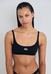 Top bikini nero accorciato realizzato in tessuto morbido, con sottili spalline e un piccolo logo metallico al centro. Il design è minimalista.