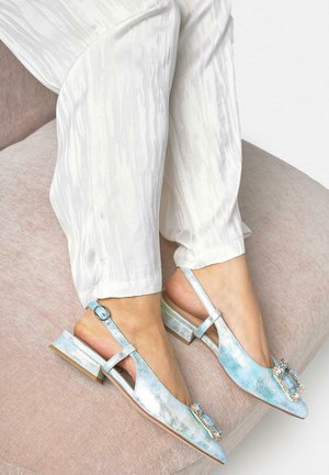 Pies con zapatos de tacón slingback metálicos azul claro con hebillas decorativas de joyas, combinados con pantalones blancos sueltos y texturizados, descansando sobre un cojín beige.