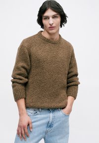 Joven con cabello oscuro de longitud media, vestido con un suéter marrón texturizado y jeans de color azul claro, de pie con una mano en el bolsillo.