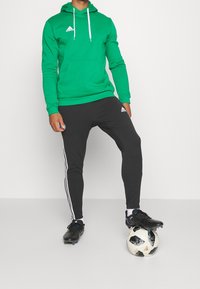 Grüne Baumwoll-Hoodie mit weißen Zugbändern, kombiniert mit schwarzen Sporthosen mit weißen Streifen. Es sind schwarze Fußballschuhe und ein Fußball vorhanden.