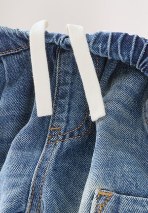 Nærbillede af denimstof, der viser en blå farve med udvaskede områder, med en hvid snor og synlige syningsdetaljer.