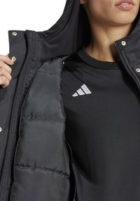 Schwarze Daunenjacke mit Kapuze, die eine glatte, glänzende Außenschicht und ein weiches, isolierendes Innenfutter aufweist. Schwarzes Shirt mit sichtbarem weißen Adidas-Logo.