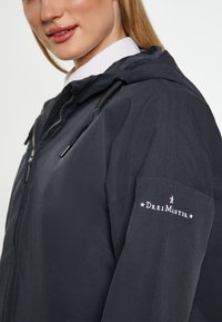 Sort vandtæt jakke med hætte, der har en frontlynlås og justerbare snore. Hvidt logo på venstre ærme.