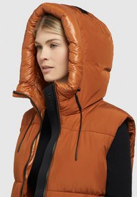Gilet imbottito in arancio bruciato con un grande cappuccio imbottito, chiusura con zip e dettagli neri. Struttura liscia e lucida con motivo trapuntato.