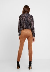 Femme aux longs cheveux bruns portant un chemisier imprimé léopard noir et brun, un pantalon en daim marron taille haute, et des bottines noires à talons hauts.