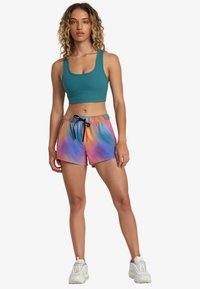 Top corto acanalado en color teal combinado con pantalones cortos de tirantes elásticos con estampado de tie-dye en tonos rosa, naranja y azul. La modelo lleva zapatillas blancas de suela gruesa.