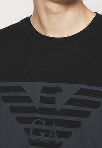 Camiseta de algodón negra con un gran logo de águila gris oscuro con rayas horizontales. Diseño clásico de cuello redondo. Textura suave.