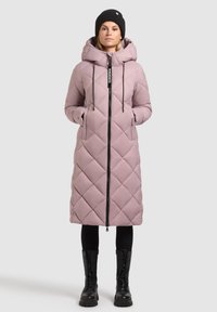 Cappotto lungo trapuntato in rosa tenue con chiusura a zip, cappuccio e dettagli neri. Presenta tasche laterali e una vestibilità rilassata. Indossato con stivali neri.
