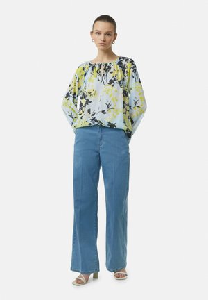 Bluse mit Blumenmuster in Hellblau, Gelb und Schwarz mit gerafftem Ausschnitt, kombiniert mit weit geschnittenen blauen Jeans. Model trägt Riemchensandalen.