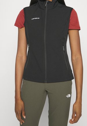 Kobieta ubrana w czarną bezrękawnik na zamek błyskawiczny, na czerwonej koszulce z krótkim rękawem oraz oliwkowe spodnie z logo na kamizelce i spodniach.