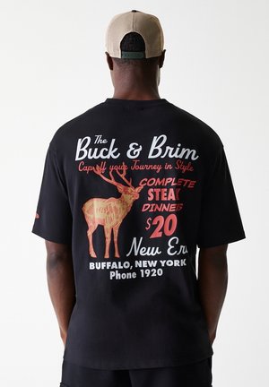 Mężczyzna w czarnej koszulce i beżowej czapce, z grafiką łosia i tekstem reklamującym obiad ze stekiem za 20 dolarów w Buffalo, Nowy Jork.