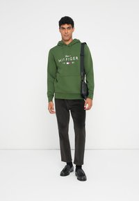 Sweat-shirt à capuche vert avec logo en texte blanc, poche kangourou et cordon de serrage. Associé à un pantalon en velours côtelé marron et des mocassins noirs. Sac en cuir sur l'épaule.