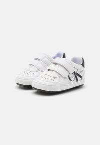 Białe skórzane buty dla małych dzieci z dwoma paskami na rzepy, perforowanymi detalami na czubku i czarnym akcentem na pięcie z logo.