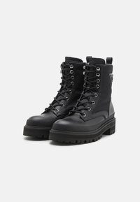 Bottes de combat noires à lacets avec une semelle épaisse, en cuir lisse et en caoutchouc, avec des accessoires en métal argenté et une étiquette de marque sur le côté.