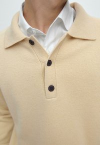 Maglione in maglia beige con scollatura a V e colletto, dotato di tre bottoni scuri e texture a coste. Indossato sopra una camicia bianca a righe.