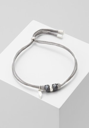 Graues verstellbares Armband mit zwei schwarzen facettierten Perlen, einer silbernen Perle und einem kleinen silbernen Blattanhänger auf weißer Oberfläche.