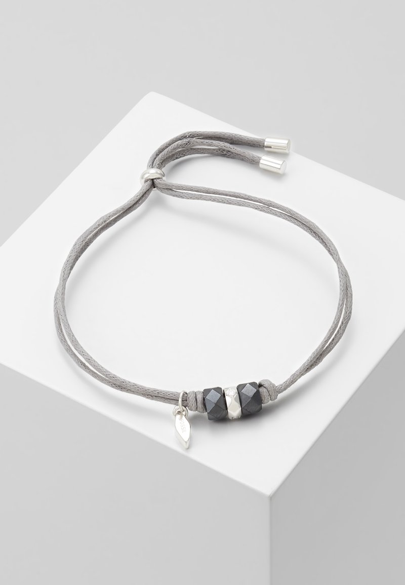 Bracelet à cordon gris ajustable avec deux perles noires facettées, une perle argentée et un petit pendentif feuille argenté sur une surface blanche.