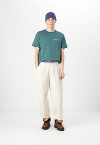 Camiseta de color azul verdoso con logo, pantalones blancos con cordón y botas marrones. Gorra de canalé azul. Diseño sencillo con corte relajado y tejidos ligeros.