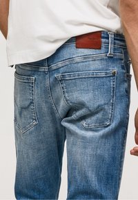 Hombre con jeans azules desgastados que tienen dos bolsillos traseros y una etiqueta de cuero marrón de la marca, combinado con una camisa blanca.