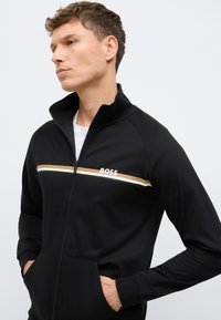 Μαύρο μπουφάν με φερμουάρ και ψηλό γιακά, με λευκές και χρυσές οριζόντιες ρίγες και λογότυπο "BOSS" στο μπροστινό μέρος. Μαλακό ύφασμα και διπλές πλαϊνές τσέπες.