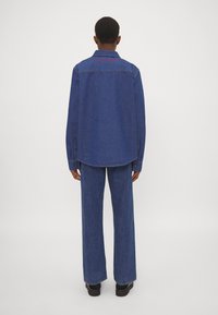 Camicia e pantaloni in denim blu, con una vestibilità comoda, maniche lunghe e orlo curvo. La camicia presenta ricami rossi sul retro.